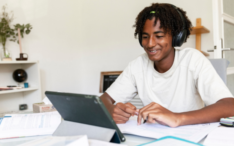 A person sitting at a desk with large over‑ear headphones on, looking toward a tablet propped up on a stand. The desk is covered with papers, notebooks, and folders. A shelf, a globe, and various items are visible in the background of the room.
